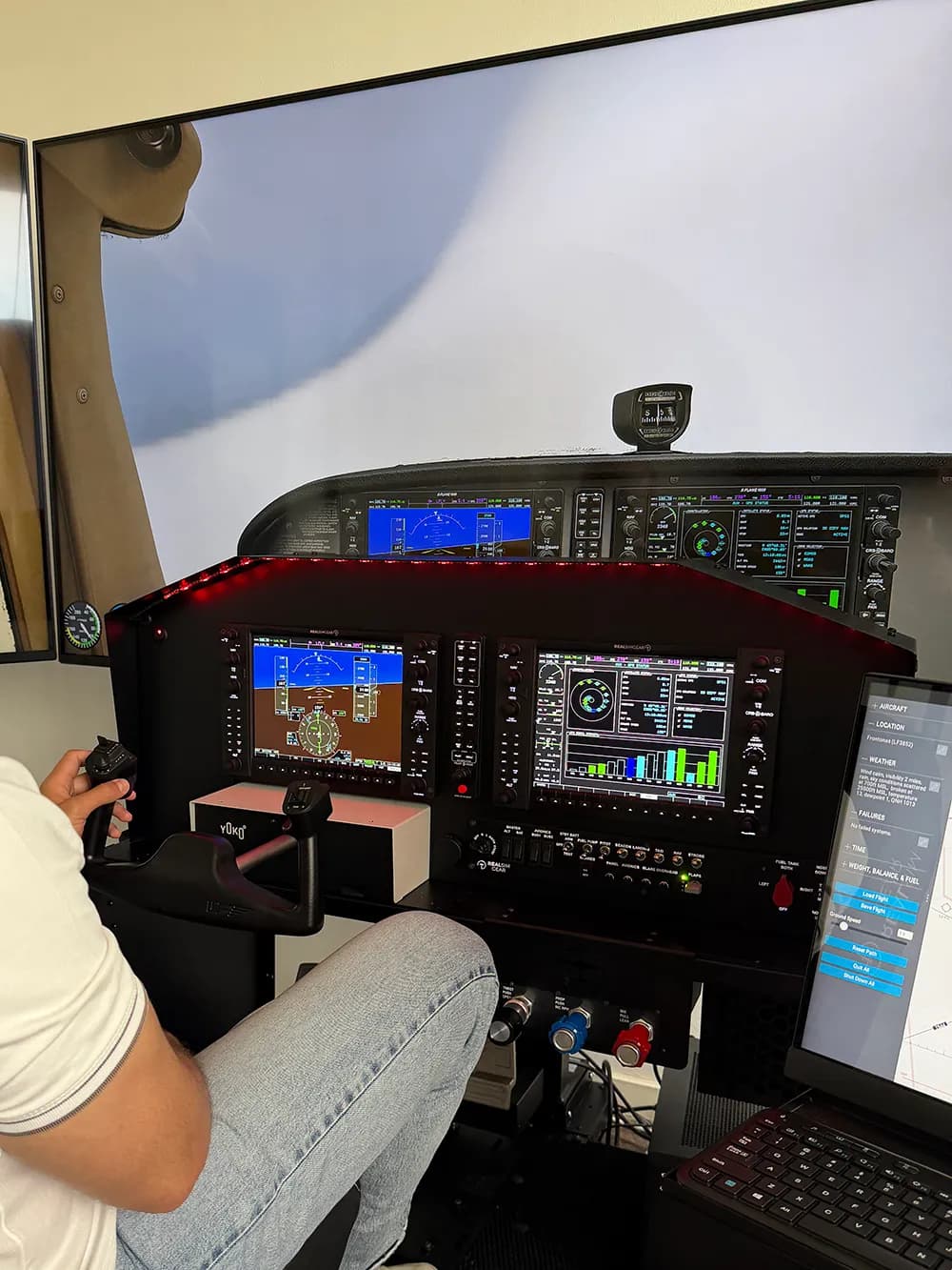 Un pilote aux commandes du simulateur, panneau G1000 et vue extérieure sur les écrans